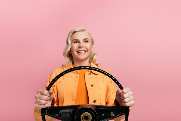 Smiling woman holding steering wheel and looking away isolated on pink  - Foto, Imagen