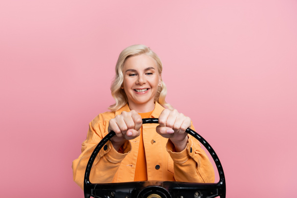 Positive blonde driver looking at steering wheel isolated on pink  - Foto, immagini