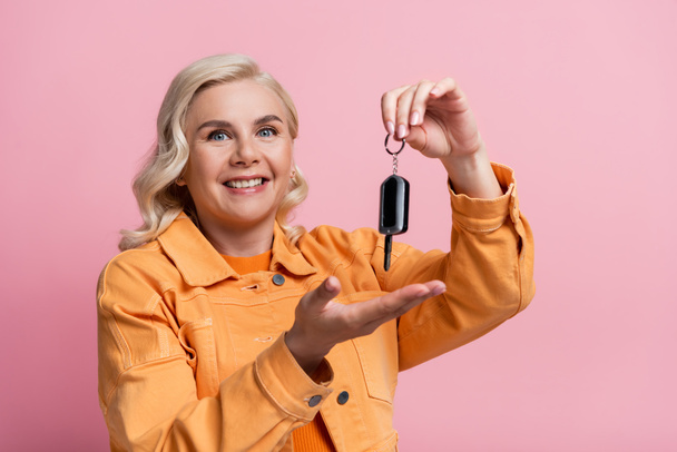 Positive blonde woman holding car key isolated on pink  - Фото, изображение