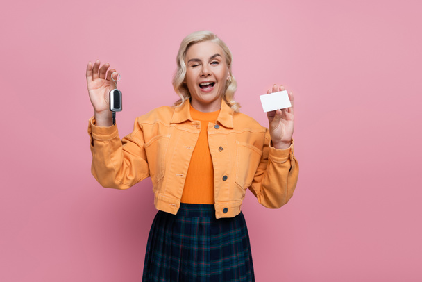 Blonde woman holding car key and driving license and winking at camera isolated on pink  - Zdjęcie, obraz