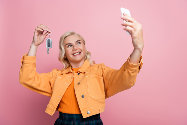 Cheerful woman holding car key and taking selfie on smartphone isolated on pink  - Foto, afbeelding