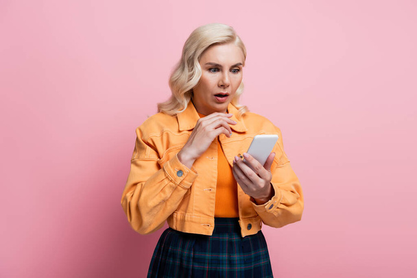 Shocked woman in orange jacket using smartphone isolated on pink  - 写真・画像
