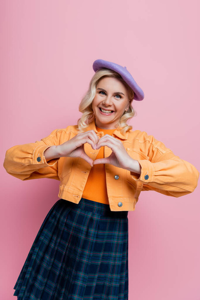 Positive blonde woman in beret showing heart sign isolated on pink  - Photo, image