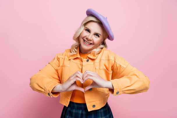 Smiling blonde woman in beret and jacket showing love sign isolated on pink  - Foto, afbeelding