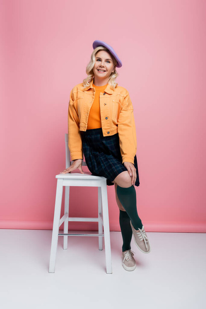 Smiling woman in skirt and beret posing near chair on pink background - Фото, изображение