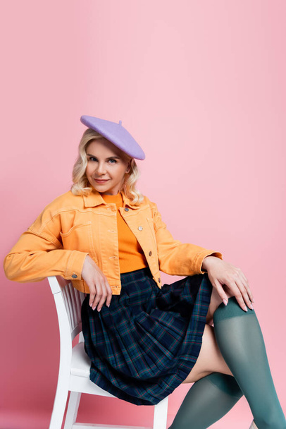 Trendy young woman in beret sitting on chair on pink background - Φωτογραφία, εικόνα