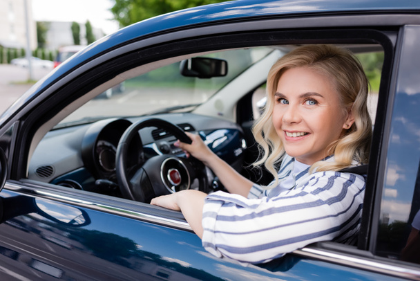 Cheerful blonde woman looking at camera while driving auto during course  - Photo, Image