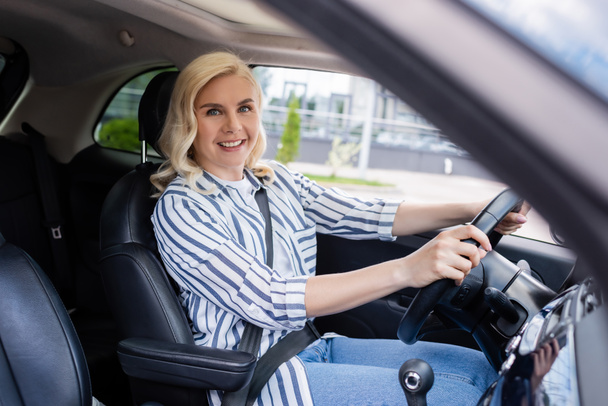 Cheerful blonde driver holding steering wheel and looking at camera in auto  - Foto, Imagen