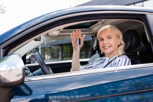 Positive blonde woman holding key while sitting in car  - Foto, Imagen