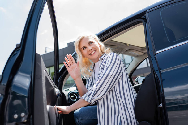 Smiling blonde woman waving hand while opening door of car outdoors  - Photo, Image