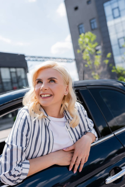 Positive blonde woman looking up from window of car outdoors  - Photo, Image