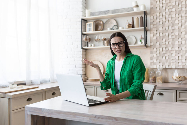 Portrait of woman at home, annoyed brunette in green shirt and glasses looking at camera and arguing, working in kitchen with laptop - Фото, изображение