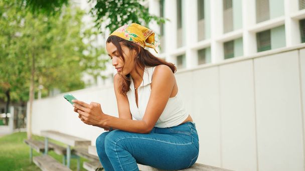 Profile of cute tanned woman with long brown hair wearing white top and yellow bandana seated using cellphone on bench in cityscape background.  - Photo, Image