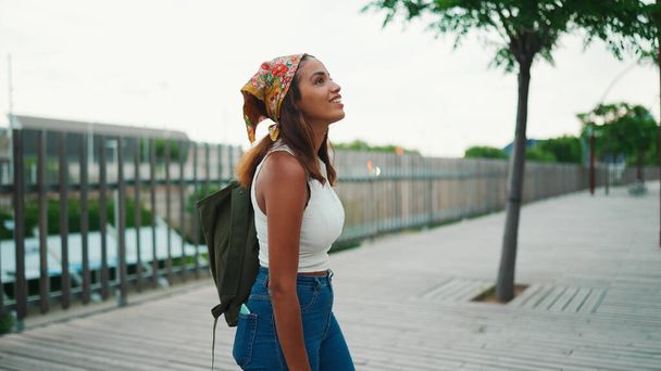 Cute tanned woman with long brown hair in white top and yellow bandana with backpack on her shoulder and looks around the modern city. - Foto, afbeelding