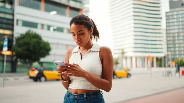 Clouse-up, cute tanned woman with ponytail wearing white top looking up at street signs and map trying to find her way using cellphone. Girl using map application outdoors - Foto, imagen