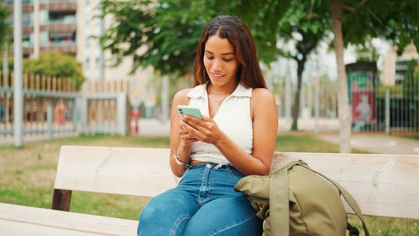 Beautiful girl with long dark hair wearing white top sits on bench and uses mobile phone. Young smiling woman writing in social networks on cellphone - Fotó, kép
