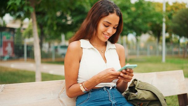 Beautiful girl with long dark hair wearing white top sits on bench and uses mobile phone. Young smiling woman writing in social networks on cellphone - Valokuva, kuva
