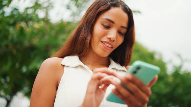 Clouse-up, beautiful girl with long dark hair wearing white top sits on bench and uses mobile phone. Young smiling woman writing in social networks on cellphone. - Foto, Bild