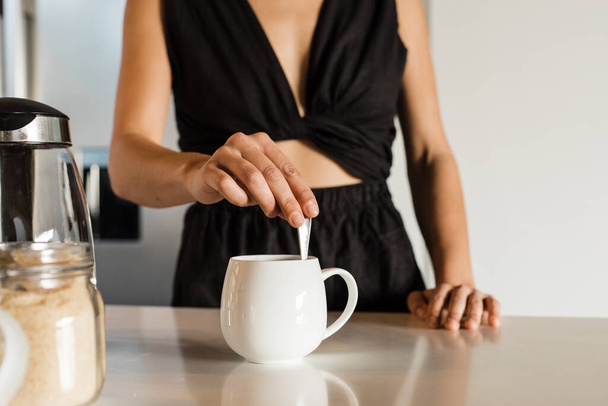 Midsection of mixed race mid adult woman wearing black attire mixing coffee in cup on kitchen island in morning - Foto, Imagem