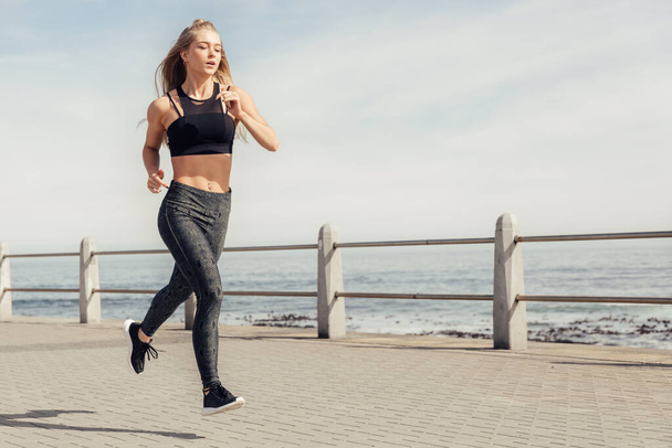 Full length of sporty caucasian young woman jogging on promenade against sea and sky during sunny day - Foto, Bild