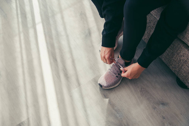 Low section of mid adult woman wearing black sports clothing tying shoelace over hardwood floor while sitting on sofa in living room - Foto, Imagem