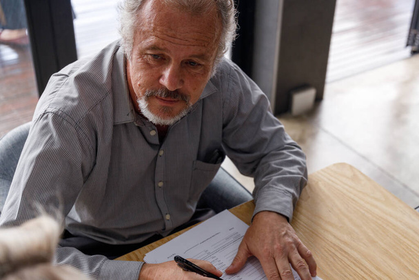 High angle view of serious caucasian mature man with job application form and pen thinking while looking away at desk in office - Fotografie, Obrázek