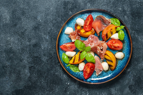 Grilled peach salad with mozzarella, prosciutto ham, red tomato, green basil and soy sauce. Fresh gourmet salad in plate on black background, close up, top view, copy space - Photo, image