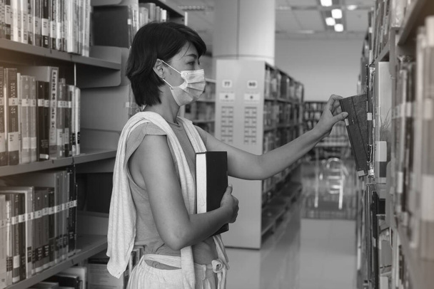 Yüzü maskeli bir kadın kütüphaneden kitabı çekiyor, siyah beyaz tonu var, bulanık ışık var. - Fotoğraf, Görsel