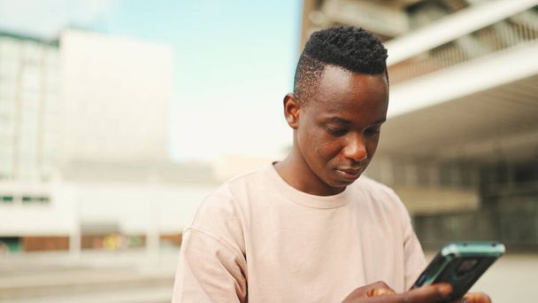 Young african student sits outside of university, uses cellphone, taps on smartphone screen - Photo, Image