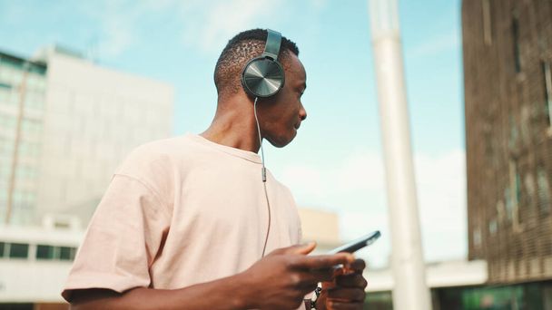 Young african student sitting outside of university wearing headphones, using phone, listening to music - Photo, Image