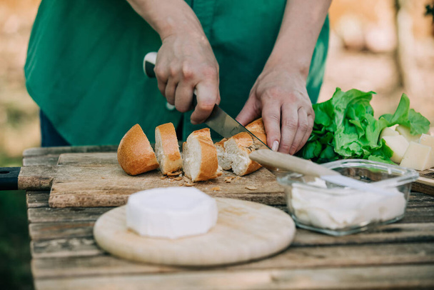 Stijlvolle vrouw in groen shirt maken een snack met brood en kaas op een tafel in de buitenlucht - Foto, afbeelding