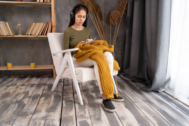woman wearing headphones knitting at home, sitting on cozy armchair in living room, happy positive female enjoying leisure time, holding needles, hobby concept - Photo, image
