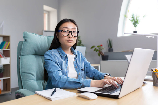 Distance Learning. A young Asian woman is a teacher, conducts online classes, lessons. He looks into the camera, explains, tells. Sitting in glasses and jeans at a table at a computer in a classroom - Φωτογραφία, εικόνα