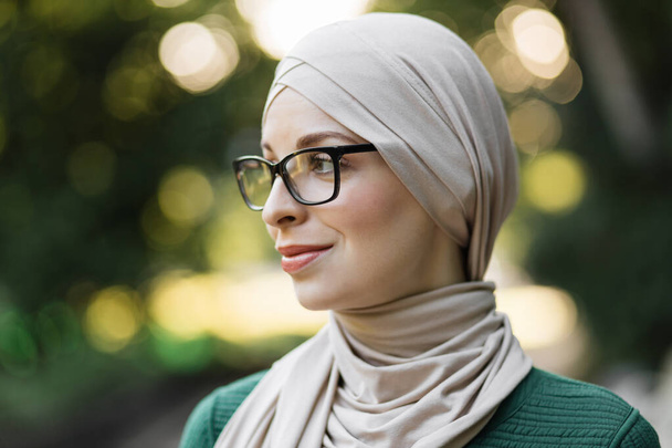 Houkutteleva hymyilevä musliminainen hijabissa katselee ulos kaupungin puiston vihreällä taustalla. Nuoren arabialaisen naisen muotokuva huivissa ja silmälasissa ulkona. - Valokuva, kuva
