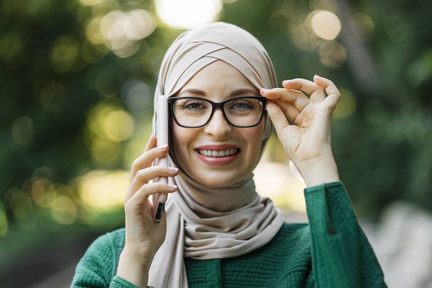 Modern stylish muslim woman in hijab and casual clothes walking in city park. Arab female talking on smartphone, taking selfie, or doing video call outdoor. - Valokuva, kuva