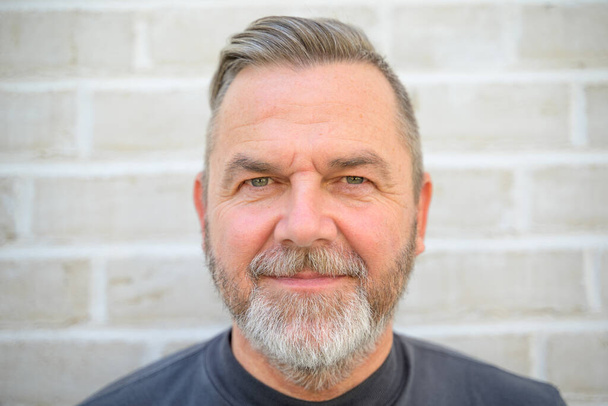 Bearded middle aged man staring intently at the camera in a close up head shot against a brick wall - Foto, imagen