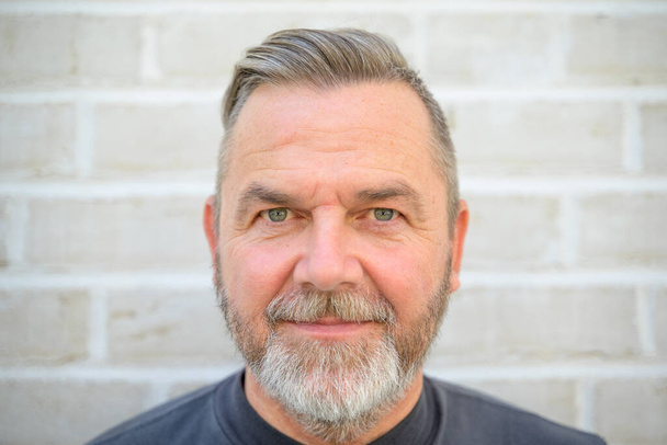 Bearded middle aged man staring intently at the camera in a close up head shot against a brick wall - Foto, Imagen