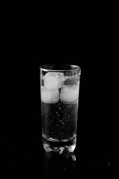Кубики льоду кидаються на склянку води зверху з бризкою на чорному тлі
 - Фото, зображення