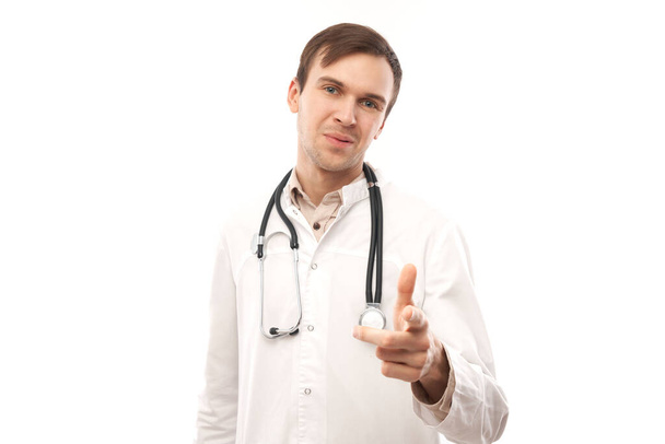 Portrait of friendly positive young male doctor in white medical coat chooses you, points finger at camera isolated on white backgroun - Foto, Bild