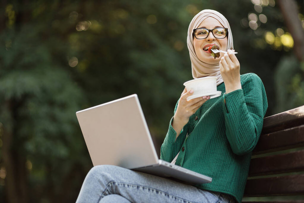 Happy smiling muslim businesswoman eating healthy salad on a break sitting on bench working on laptop. Female dieting nutrition concept. Attractive islam smiling girl enjoying veggie meal. - Zdjęcie, obraz