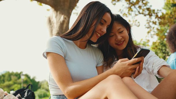 Young multi ethnic smiling women are watching videos on their phone. Girls laugh at picnic on summer day outdoors - Foto, imagen