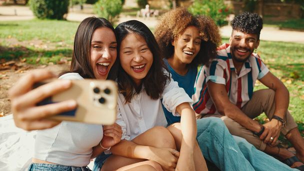 Happy multiethnic young people are looking at taking selfie on summer day outdoors. Group of smiling friends posing at smartphone camera - Foto, afbeelding