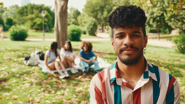 Close-up of young man with curly hair wearing striped shirt sitting in park having picnic on summer day outdoors, smiling and talking with friends - Foto, Bild