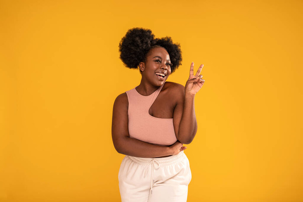 Funny young afro woman smiling to the camera, showing peace symbol. Real people emotions. A lot of copy space on the yellow studio background - Foto, imagen