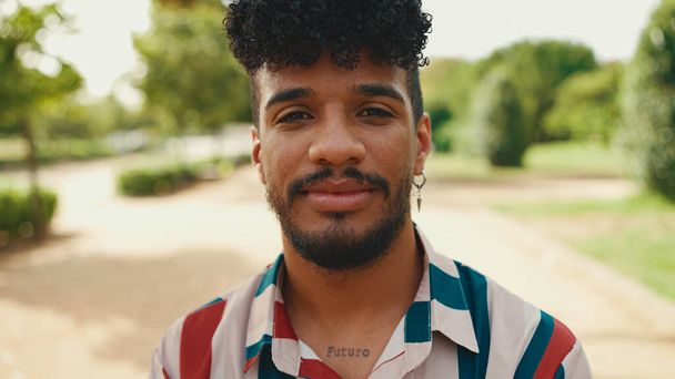Close-up of young man with curly hair wearing striped shirt sitting in park having picnic on summer day outdoors, smiling and talking with friends - 写真・画像