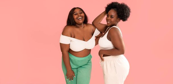 Happy overjoyed two afro women smile broadly, having fun together, posing on pink pastel studio background. Body positive concept - Foto, Bild