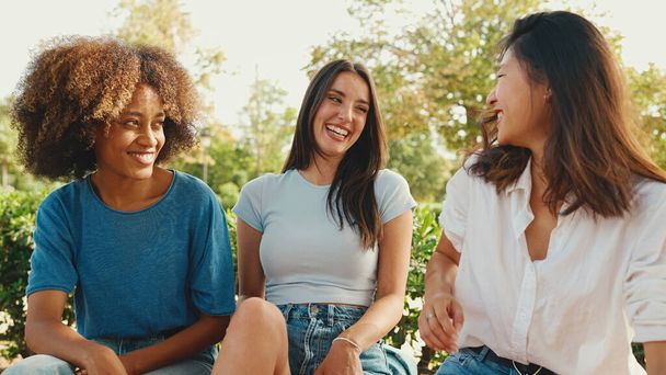 Happy multiethnic young women talking while sitting on park bench on summer day outdoors. Group of girls talking and laughing merrily in city park - Foto, imagen