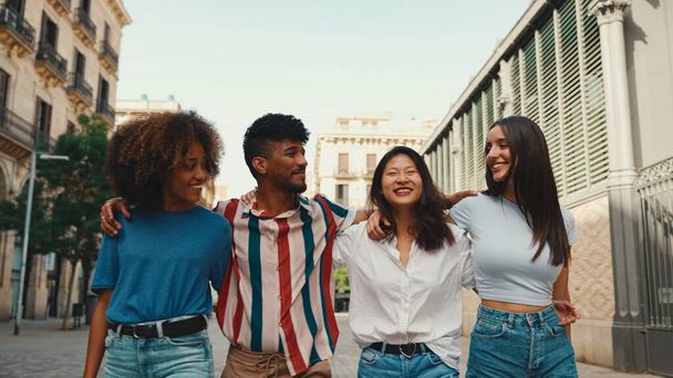 Happy multiethnic young people walk embracing on summer day outdoors. Group of friends are talking and laughing merrily while walking along on the street - Zdjęcie, obraz