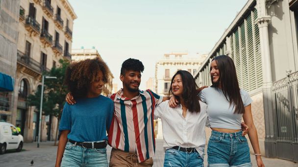 Happy multiethnic young people walk embracing on summer day outdoors. Group of friends are talking and laughing merrily while walking along on the street - 写真・画像