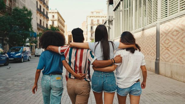 Happy multiethnic young people walk embracing on summer day outdoors. Group of friends are talking and laughing merrily while walking along on the street - Foto, imagen